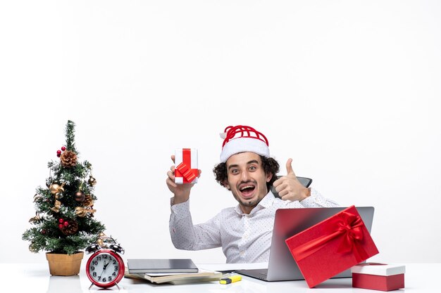 산타 클로스 모자는 사무실에 앉아 흰색 배경에 그의 선물을 들고 웃는 만족 된 젊은 사업가와 함께 새해 분위기