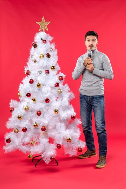 Новогоднее настроение с удивленным позитивным парнем в джинсах, стоящим возле украшенной елки на красном