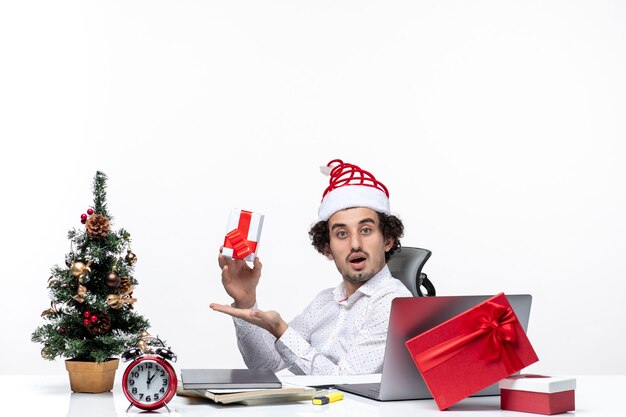 New year mood with surprised young businessman with santa claus hat sitting in the office and holding his gift posing for camera on white background