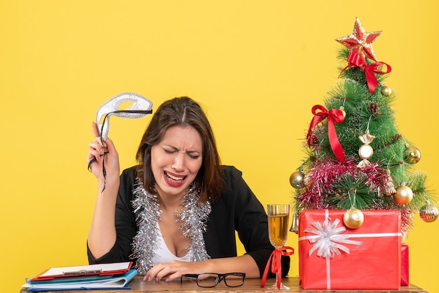 Бесплатное фото Новогоднее настроение с задумчивой очаровательной дамой в костюме, держащей маску и плачущей в офисе на желтом изолированном