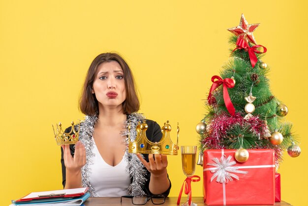 New year mood with thoughtful charming lady in suit holding crowns in the office on yellow isolated 