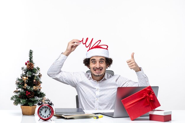 New year mood with unsure uncertain young businessman touching his funny santa claus hat pointing above in the office on white background