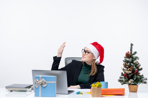 New year mood with young attractive woman with a santa claus hat sitting at a table with a Christmas tree and a gift on it saying hello in the office