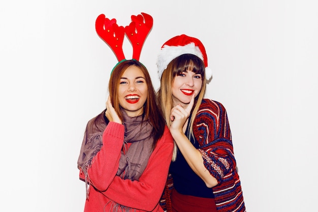 Free photo new year party. two beautiful girls in funny masquerade santaâs hats send kiss . indoor stockpile image of best friends posing. isolate.