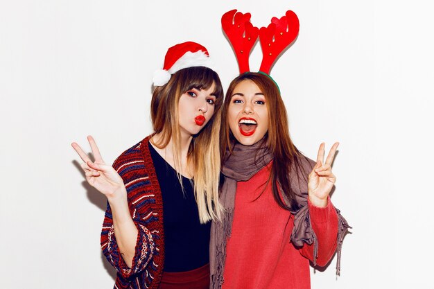 새해 파티. 재미있는 가장 무도회 산타 모자를 쓴 두 명의 아름다운 소녀가 키스를 보냅니다. 가장 친한 친구 포즈의 실내 비축 이미지입니다. 격리.