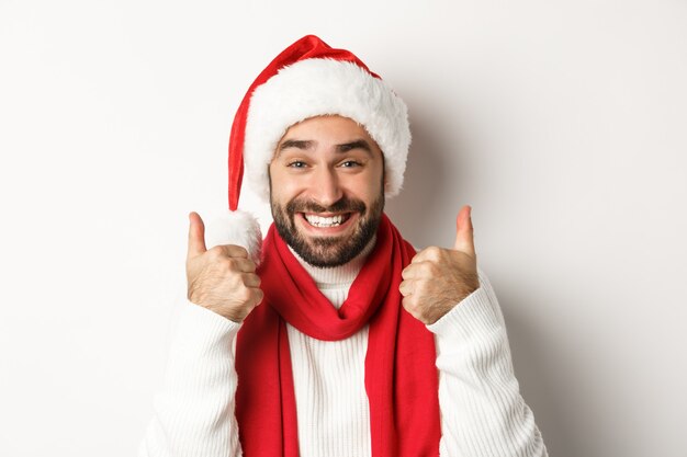 신년 파티와 겨울 휴가 개념입니다. 흰색 배경 위에 서 있는 승인, 좋아요 및 동의에 엄지손가락을 보여주는 산타 모자를 쓴 행복한 남자의 클로즈업