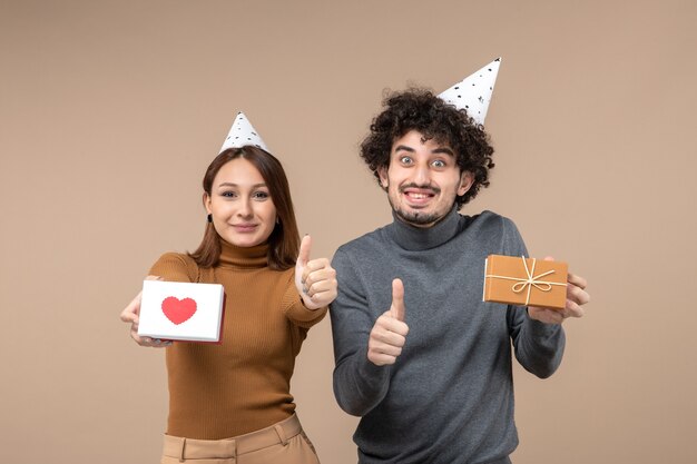 젊은 부부와 함께 새해 촬영은 마음으로 새해 모자 소녀와 선물을 가진 사람을 착용하고 회색에 확인 제스처를 만듭니다.