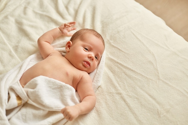 샤워 후 신생아는 흰색 담요에 침대에 누워 수건에 싸여 있고, 멀리 보이는 유아, 욕실 후 부드러운 피부를 가진 매력적인 아이, 편안한 아이.