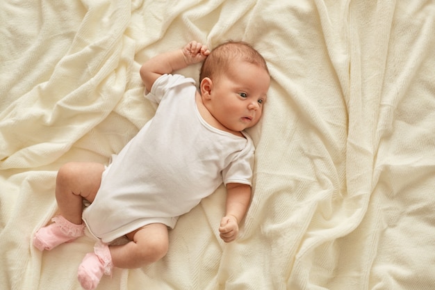 Newborn baby girl or boy lying on blanket on bed looking away, wearing white bodysuit and socks, infant studying world around, has sleepy expression.