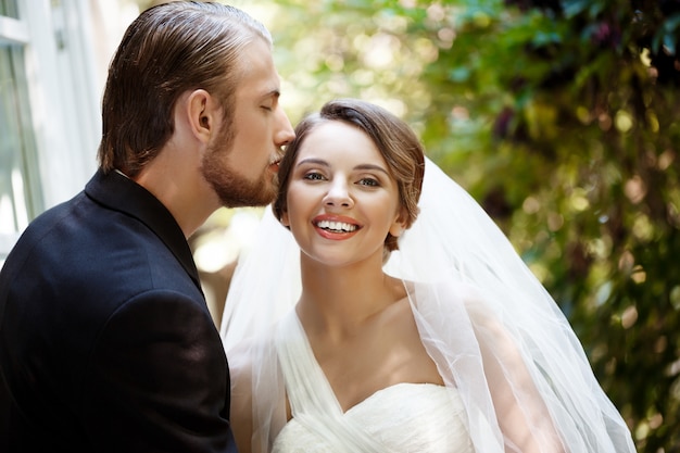 Newlyweds in suit and wedding dress smiling, kissing in park.