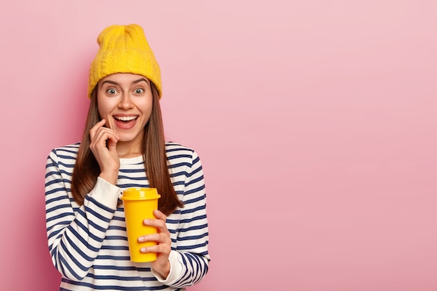Милая очаровательная женщина держит бумажный стаканчик с кофе, носит желтую шляпу и полосатый свитер, широко улыбается, имеет естественную красоту, изолированную на розовой стене