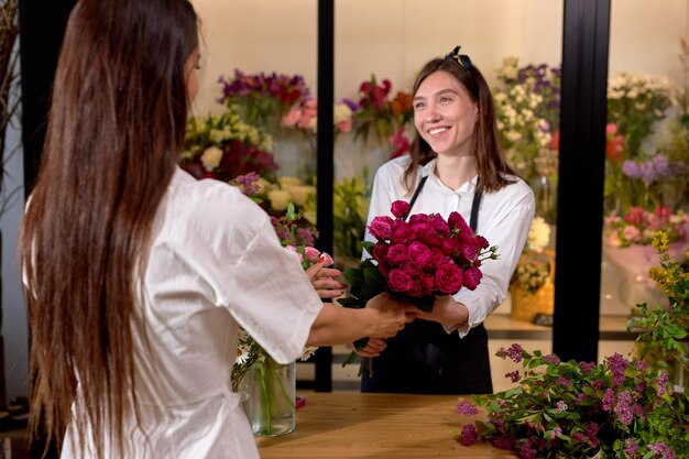 素敵な​白人​女性​の​花屋​が​クライアント​に​ピンク​の​バラ​の​美しく​装飾された​花束​を​与え​、​長い​髪​の​ブルネット​の​女性​が​新鮮な​花​の​最高​の​花束​を​手​に​入れる​ために​小さな​フラワー​ショップ​に​来ました