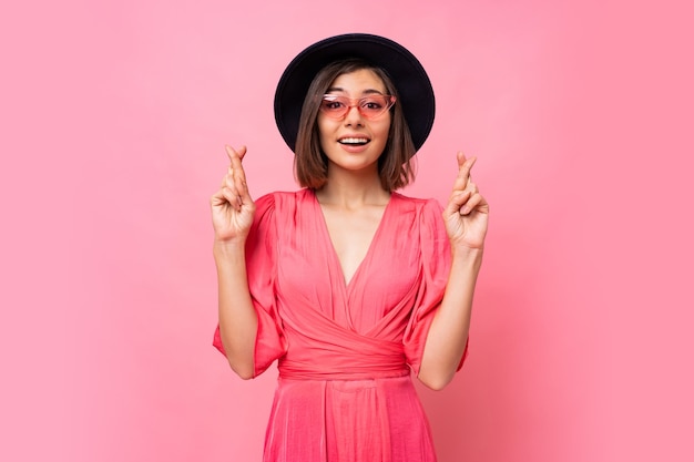 Nice cheerful woman wishes to do her best to keep her fingers crossed, has the best, wearing in dress  isolated on pink wall. People, body language concept. Copyspace.