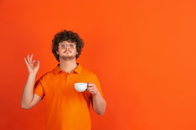 おいしいコーヒー、うれしいです。オレンジ色の壁に白人の若い男のモノクロの肖像画。カジュアルなスタイルの美しい男性の巻き毛モデル。