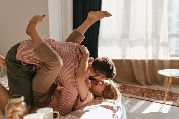 Nice couple in pastel-colored clothes is lying on bed and in good mood is fooling around in bright apartments.