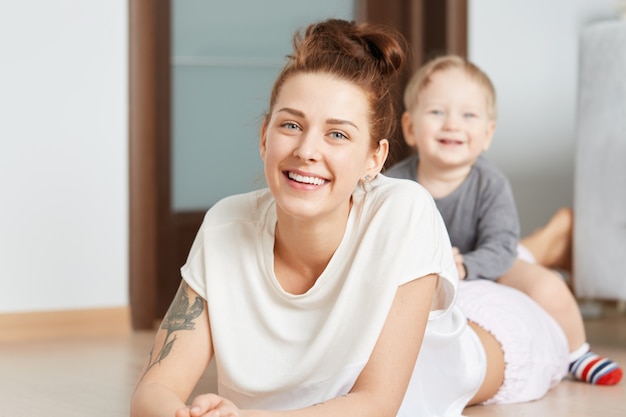 Free photo nice family shot of young mother and her little son playing on the floor at home. attractive caucasian woman in white top, lying on her belly. smiling kid straddling happily on her shinny mommy.