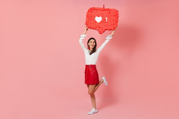 Nice girl in bright outfit smiling and joyfully demonstrating Like from social network on pink background.