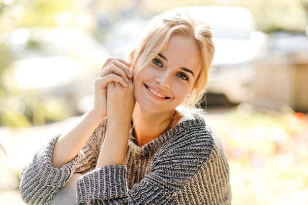 Nice girl in trendy oversized sweater leaned and looking at camera against background of cars with smile