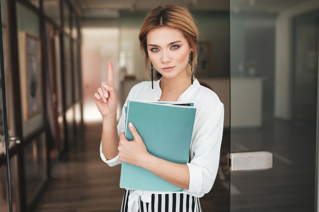 Nice girl with books in hand looking in camera and showing no gesture. Portrait of young girl with blond hair in white shirt standing near door and holding notebooks in hand.