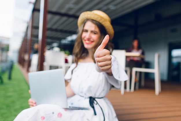 Nice girl with long hair in hat is sitting on floor on the terrace. She wears a white dress with naked shoulders, red lipstick. She has a laptop on knees. Focus on her hand  gesture on front.