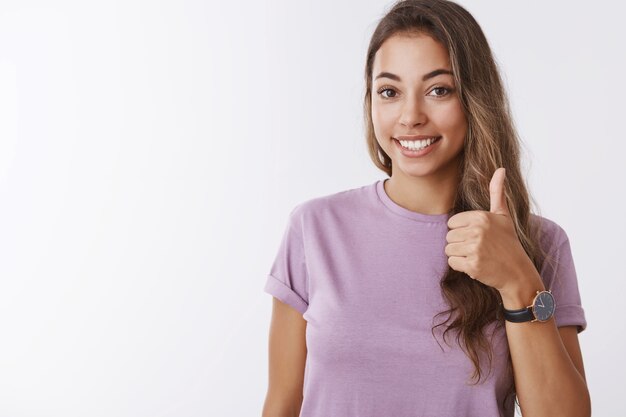 Nice I in. Friendly outgoing gorgeous young smiling girl showing thumb up saying yes, approving liking awesome interesting plan, grinning accepting terms, no problems, standing white wall