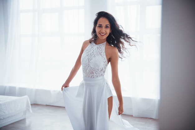 Nice lighting. Beautiful woman in white dress stands in white room with daylight through the windows