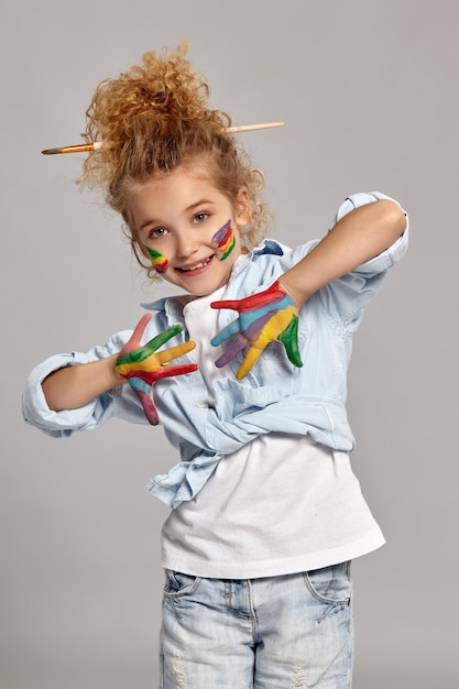 Nice little girl having a brush in her chic haircut, wearing in a blue shirt and white t-shirt. She is posing with a painted arms and cheeks, looking at the camera and smiling, on a gray background.