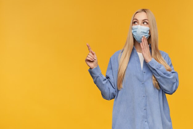 Nice looking woman, beautiful girl with long blond hair. wearing blue shirt and medical face mask. watching at the camera and pointing to the left at copy space, isolated over orange background