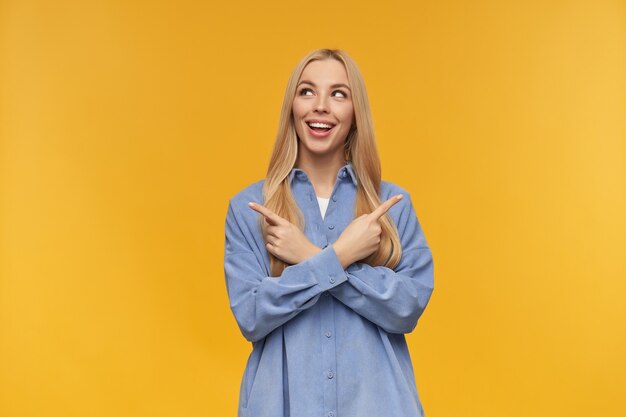 Nice looking woman, beautiful girl with long blond hair. wearing blue shirt. people and emotion concept. watching to the left and pointing both sides at copy space, isolated over orange background