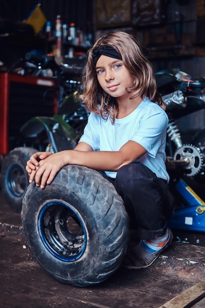 Nice small girl is posing for photographer with big wheel from car at auto service.