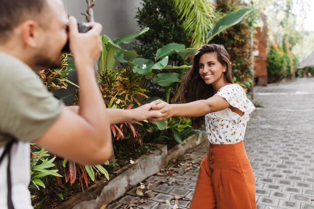 Nice tanned lady in orange pants holding her boyfriends hand Man photographs girl on path with tropical plants