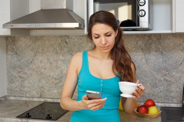 A nice woman in a green t-shirt and dark hair is standing in the kitchen with a mug and a phone in her hand.