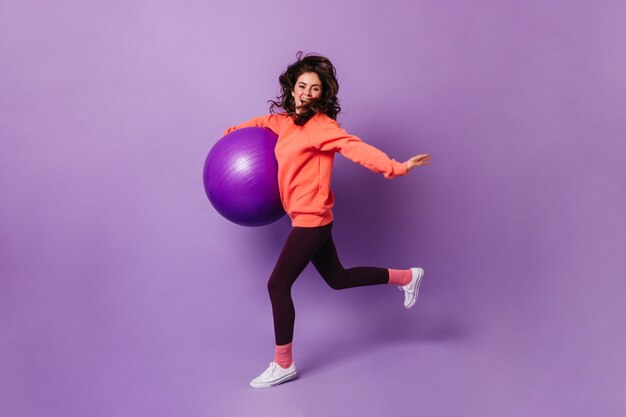 Nice woman in pink socks, orange sweatshirt and dark leggings runs on purple wall with fitball