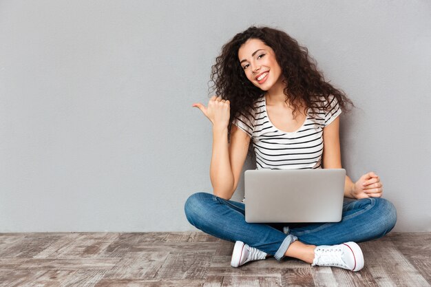 Nice woman with beautiful smile sitting in lotus pose on the floor with silver computer on legs gesturing thumb aside submitting something copy space