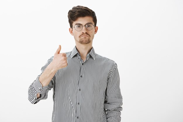 Nice work man. Pleased impressed attractive male student in glasses, showing thumbs up and sucking lips, giving approval and liking idea or project of friend, sharing his opinion over gray wall