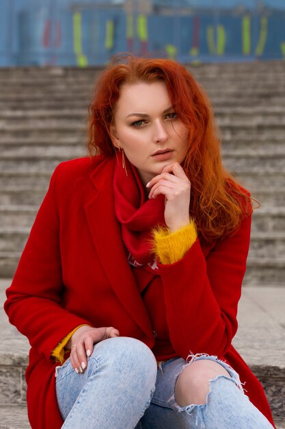 A nice young woman with red hair in a red coat and jeans is sitting on the steps outside