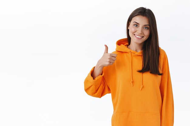 Nicely done. Pleased happy smiling caucasian female in orange hoodie, tilt head and look satisfied, show thumb-up to express her positive opinion