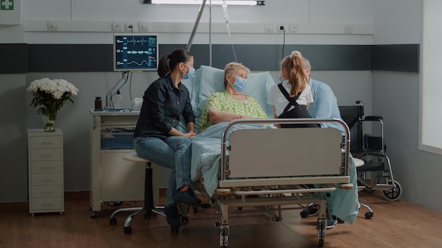 Niece visiting senior patient with face mask in hospital ward with mother and old man. Sick woman enjoying visit from daughter and child, giving comfort to cure disease during pandemic.