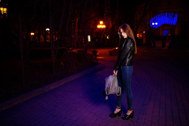 Ночной портрет девушки-модели в очках, джинсах и кожаной куртке с рюкзаком в руках на фоне гирлянды синих огней городской улицы