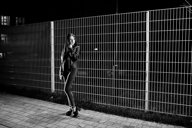 Night portrait of girl model wear on jeans and leather jacket against iron fence