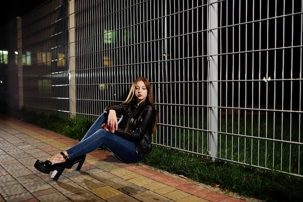 Ночной портрет девушки-модели в джинсах и кожаной куртке на фоне железного забора