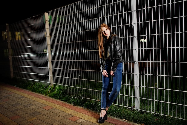Night portrait of girl model wear on jeans and leather jacket against iron fence