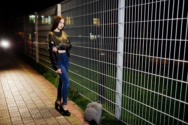 Ночной портрет девушки-модели в джинсах и кожаной куртке на фоне железного забора