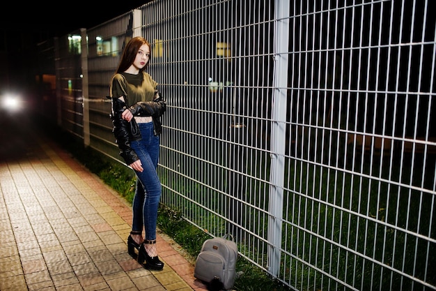 Night portrait of girl model wear on jeans and leather jacket against iron fence