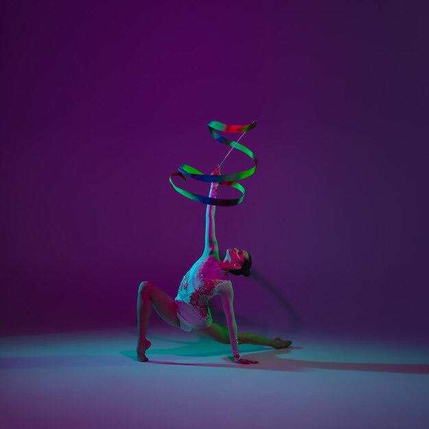 Ночь. Молодая спортсменка, артистка художественной гимнастики танцует, тренируется с лентой, изолированной на фиолетовом студийном фоне в неоновом свете. Красивая девушка практикуется с оборудованием. Грация в исполнении.