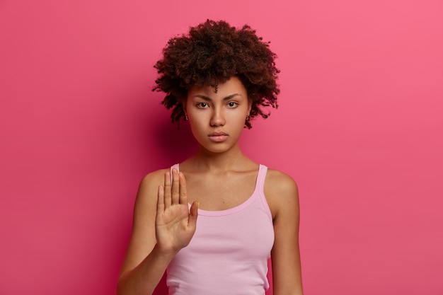 No, I dont want. Serious looking curly young woman makes stop gesture with assertive expression, rejects bad offer, wears casual vest, looks confidently , isolated on pink wall