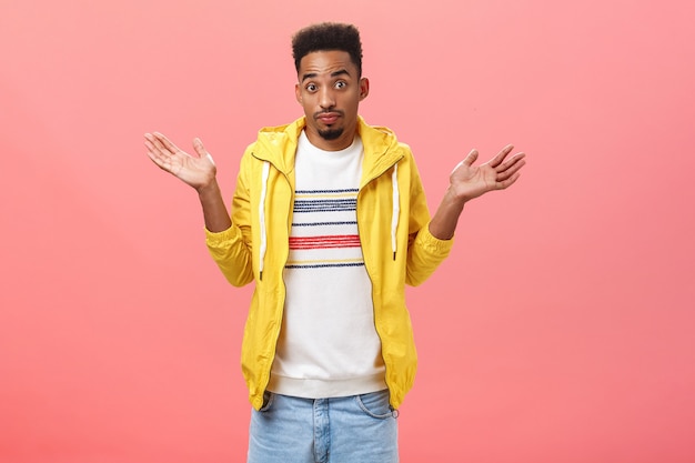 No idea bro. Unsure clueless african american guy with beard and afro hairstyle in yellow trendy jacket shrugging with palms raised near shoulders being uncertain and unaware over pink wall.