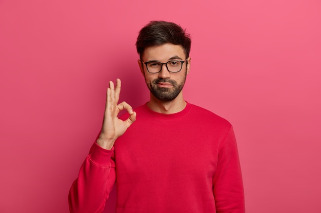 No problem concept. bearded man makes okay gesture, has everything under control, all fine gesture, wears spectacles and jumper, poses against pink wall, says i got this, guarantees something Free Photo