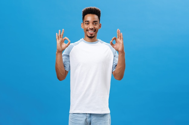 No problem. Portrait of joyful optimistic dark-skinned boyfriend with cool haircut in casual t-shirt showing okay gesture liking great idea of friend smiling delighted posing over blue background