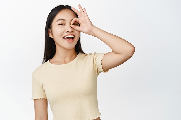 No problem Smiling happy asian woman showing zero okay gesture on eye laughing carefree standing over white background